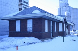 Single-story brick building with a dark roof and white-trim windows in snowy surroundings near industrial silos.